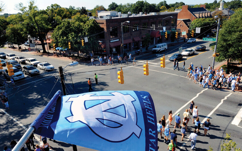 Aerial view of a downtown street crossing on a sunny day with a UNC Tar Heels flag flluttering close up