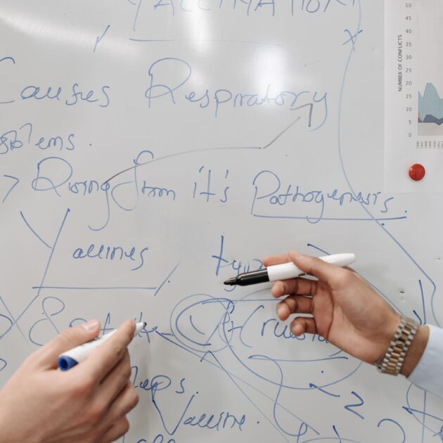 Hands holding markers writing notes on a whiteboard about COVID-19 and vaccination.