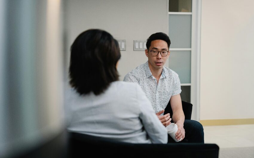 Two young professionals sitting in conversation in office setting, young Asian man faces camera