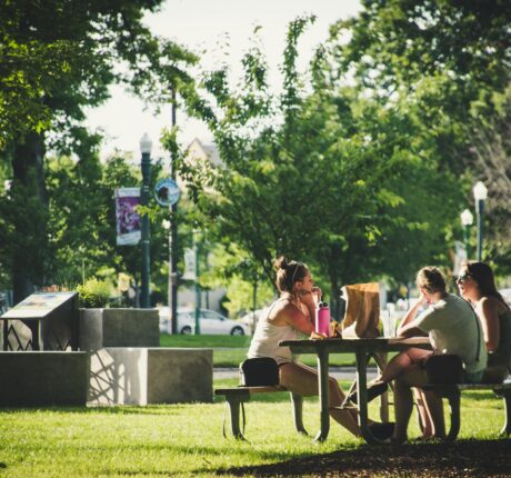 Three people enjoying a meal outside in the sun on a park bench