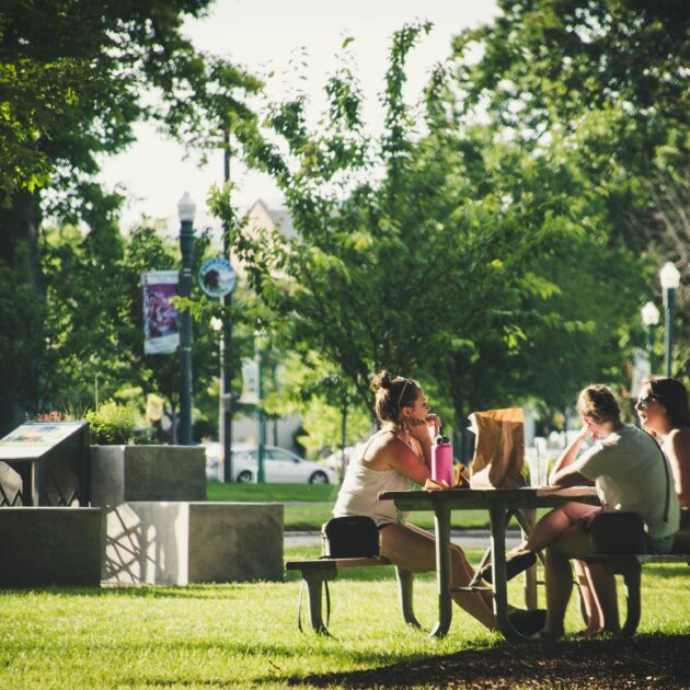 Three people enjoying a meal outside in the sun on a park bench