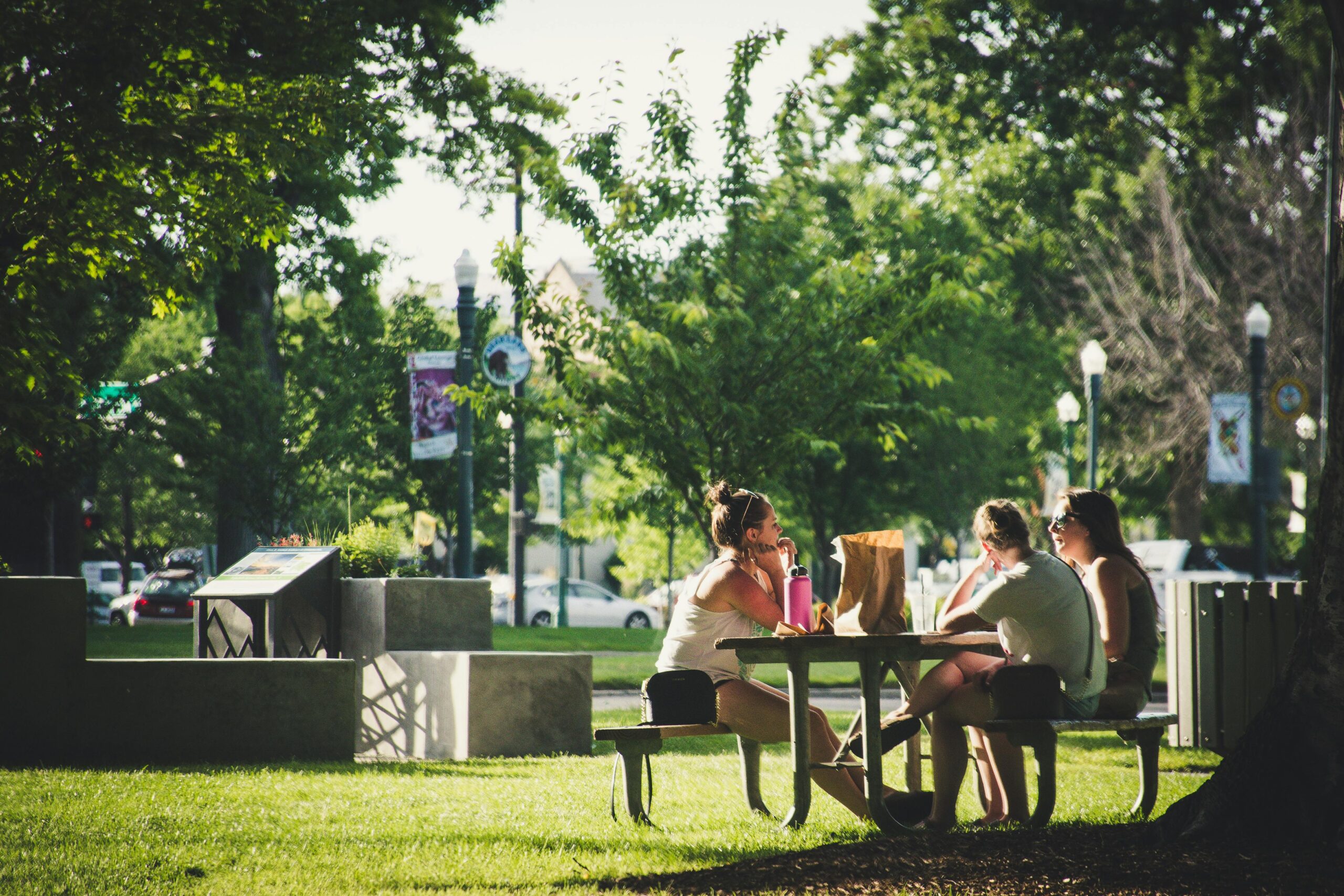 Three people enjoying a meal outside in the sun on a park bench
